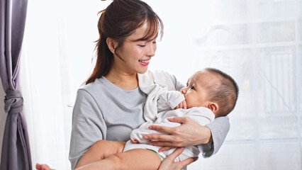 Obraz premium Young happy asian mother holding adorable infant baby near window in bright room showing love care motherhood tenderness and family bond in domestic lifestyle