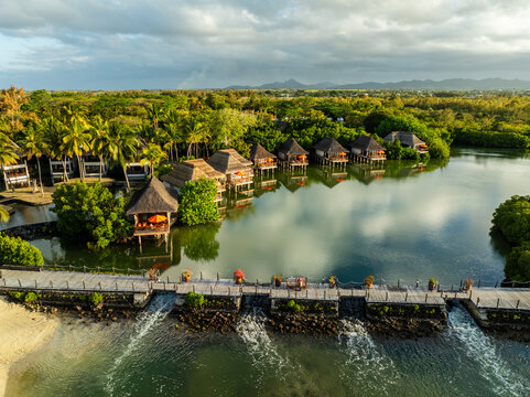 Aerial view of thatched-roof bungalows standing above the water, reflecting the sky, surrounded by lush greenery, Prince Maurice, Flacq District, Mauritius.