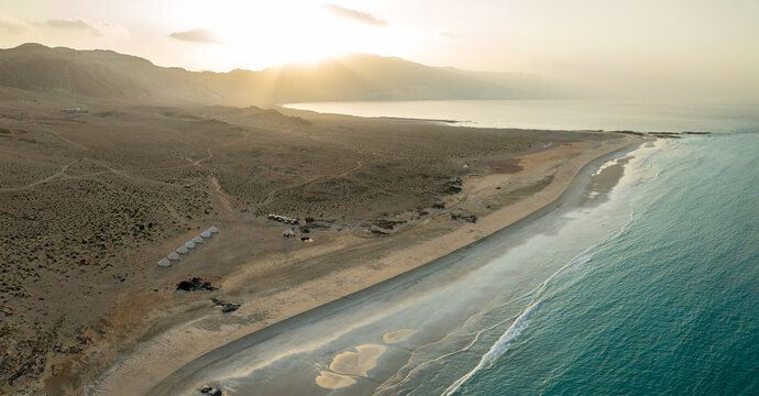 Aerial view of the meeting between arid desert terrain and the turquoise sea near a cluster of tents, Erisel, Socotra.