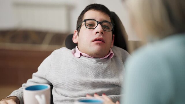 Young man with a physical condition sitting in a wheelchair and having an animated conversation with a woman while drinking from a mug