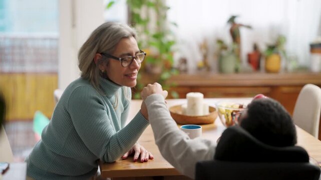 Devoted mother and her son with a physical condition sit at the dining table. The son, who uses a wheelchair, and his mother talk and hold hands