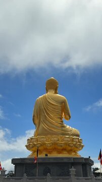 Vertical close view of a golden Buddha statue with Vitarka mudra raised hand, set against open sky and drifting clouds above the temple grounds. Buddhist gesture.