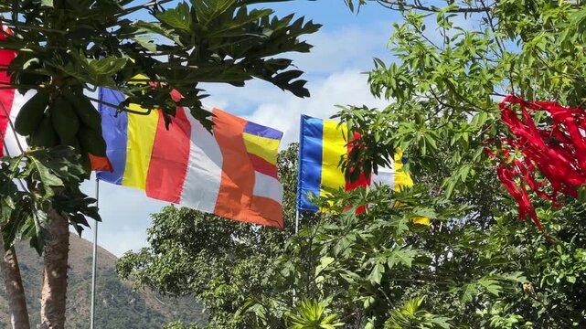 Several Buddhist flags wave together surrounded by trees under a blue sky near a temple or spiritual location. Religious celebration.