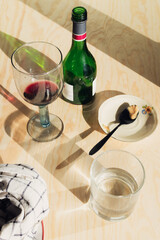 Top view of a glass of red wine and a glass of water on a wooden table with a green bottle and a small saucer in bright natural light
