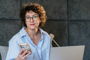 Fototapeta premium Confident White woman in her 40s with glasses holding smartphone beside laptop in bedroom