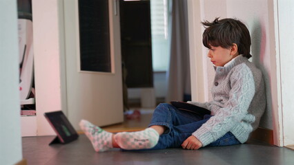 Fototapeta premium Young boy sitting on floor, eating bread, watching tablet, relaxed and engaged, casual home setting, child enjoying snack, focused expression, indoor activity