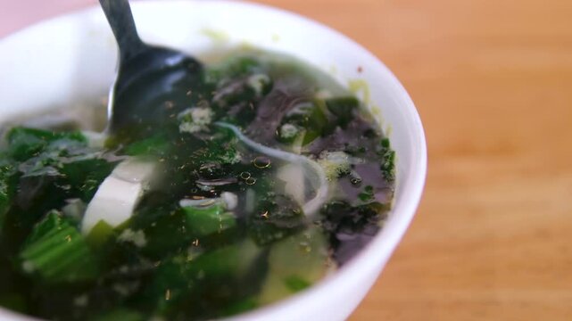miso soup Hand uses spoon to lift pork ball and seaweed from yellow bowl of Thai soup on wooden table, under warm indoor lighting with shallow depth of field