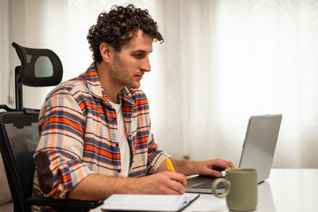 Concentrated young businessman sitting at the table, writing notes and using laptop while working at his comfortable home.	