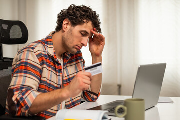 Stressed and depressed young man using laptop and credit card for online banking while sitting at his home. He is having financial problems.	
