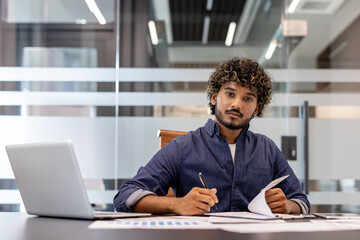 Fototapeta premium Portrait of a serious young Indian businessman working in the office with a laptop and documents, looking at the camera