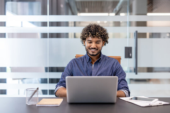 Smiling young Indian man sitting at a desk in the office and working on a laptop