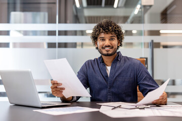 Fototapeta premium Portrait of a smiling young Indian businessman sitting in the office in front of the camera, working on a laptop and with documents