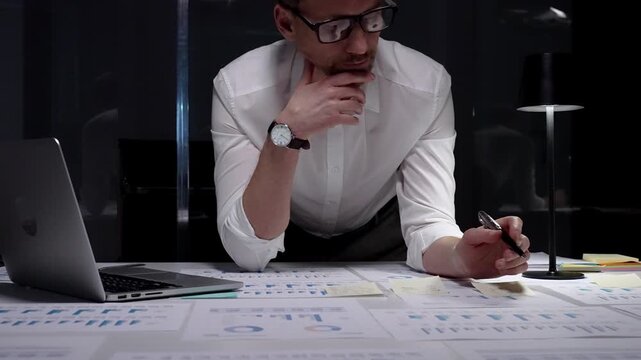 Dedicated businessman intensely analyzing financial documents and charts spread across his desk under a table lamp, concentrating on complex data in a dark office while working late evenings