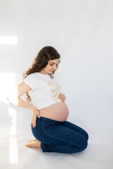 Fototapeta premium Beautiful pregnant woman in white t shirt and jeans posing in studio. Maternity tenderness parenthood new life. Happy pregnant woman on white background.