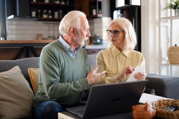 Senior couple discussing finances at home, talking seriously while reviewing documents and laptop....