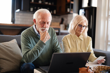 Fototapeta premium Senior couple focused on reviewing financial documents at home, using laptop and paperwork. Serious expressions reflect concern about budgeting, retirement planning, and rising expenses