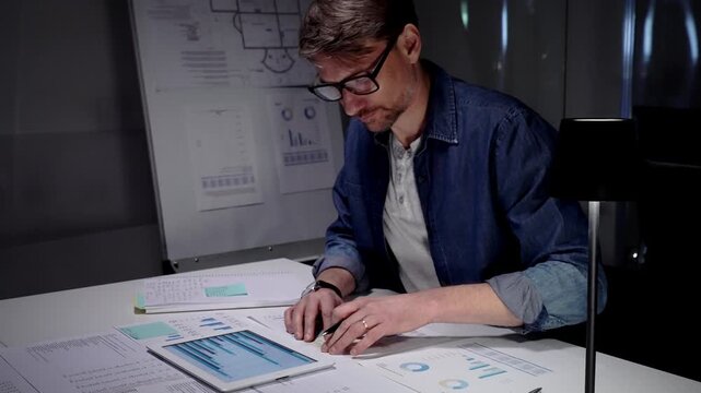 Professional man working late in a dark office, analyzing business data reports and using a digital tablet, managing overtime and workload under focused concentration