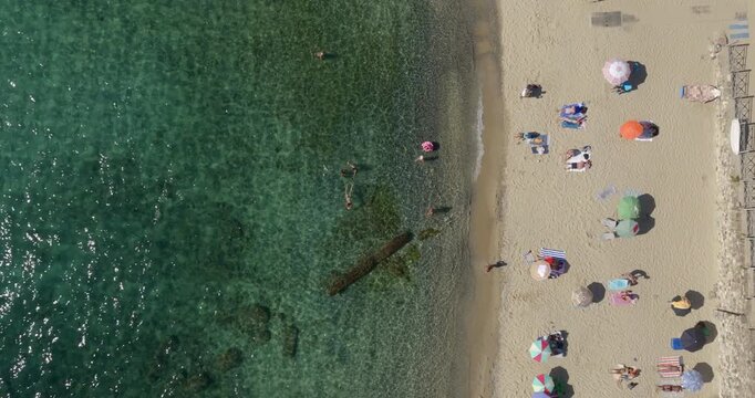 Aerial perpendicular view of a sunny Mediterranean beach. The crystal-clear turquoise water meets a golden sandy shore where colorful beach umbrellas and sunbathers are scattered across the sand.