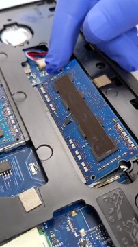 A gloved hand carefully inserts a blue RAM module into a laptop's internal circuitry for an upgrade or repair