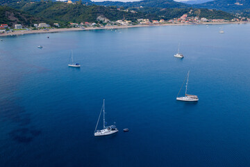 Aerial drone perspective of sailboats resting on the calm, deep blue Ionian Sea at Agios Georgios Pagon, Corfu. The tranquil scene captures the essence of a Greek island summer holiday.