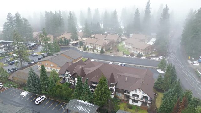 Residential buildings west of Icicle Ridge appear through dense fog along valley floor near Tumwater Canyon. Brown roofing, wet pavement, softened tree lines blend winter light in Leavenworth, WA