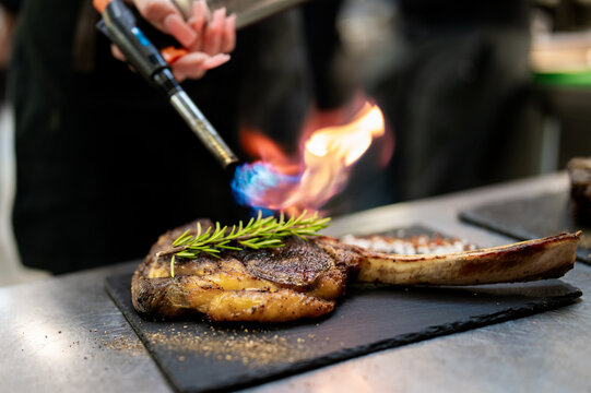 A chef uses a professional kitchen torch to sear a large tomahawk steak topped with fresh rosemary on a slate plate