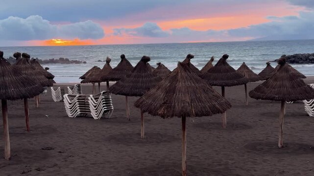 Empty beach with thatched straw parasols and stacked sun loungers at sunset by the ocean. tropical resort, summer travel,