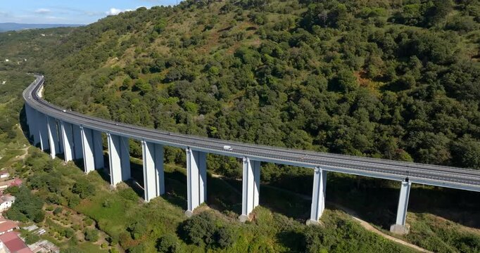 Aerial view of a modern curved highway viaduct. The long concrete structure, supported by massive pillars, gracefully cuts through a densely forested valley. A few vehicles are visible on the road.
