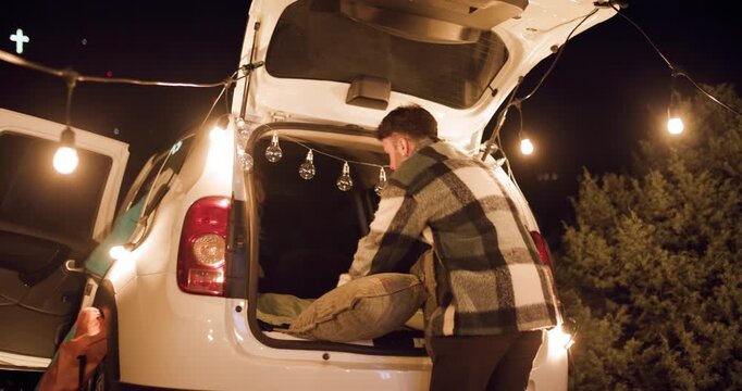 Man prepares his car for a night under the stars with friends in the forest while setting up bedding and lighting