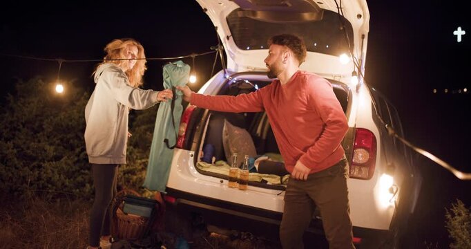 Couple prepares for camping at night in the forest under string lights with their car open and outdoor gear