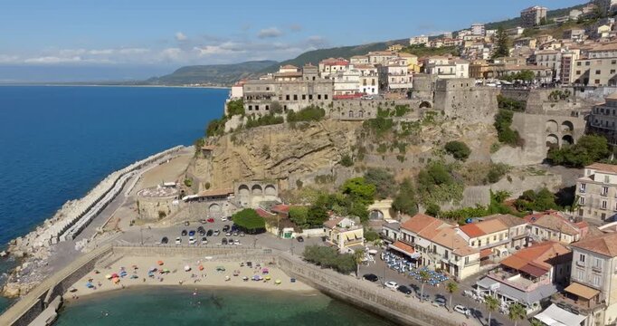 Aerial view of a small, tranquil sandy beach nestled against a sturdy stone pier and harbor wall. There are a parking area and seaside buildings border the shore. It's the urban beach of Pizzo, Italy.