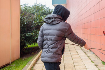 Rear view of teenage boy in hooded puffer jacket walking along urban alley holding a stick, moody city scene suggesting tension and risky behavior concept © Татьяна Швец