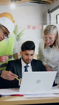 Likable successful experienced diverse male and female construction specialists at the head with responsible bearded foreman browsing bluprint or scheme of future building on laptop screen during