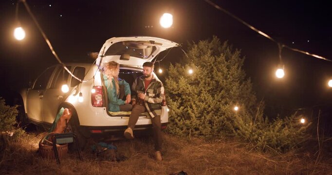 Couple enjoys evening at campsite with lights and snacks parked near trees in the mountains during autumn season