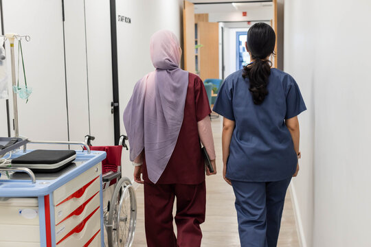 Diverse female nurses walking down clinic hall wearing scrubs with tablet past cart and wheelchair