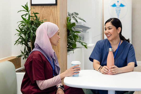 Female healthcare workers in scrubs, hijab chatting, holding cup and peach bottle in staff lounge