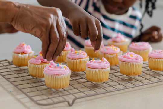 African American senior woman, striped girl sprinkling pink-vanilla cupcakes on metal rack at home