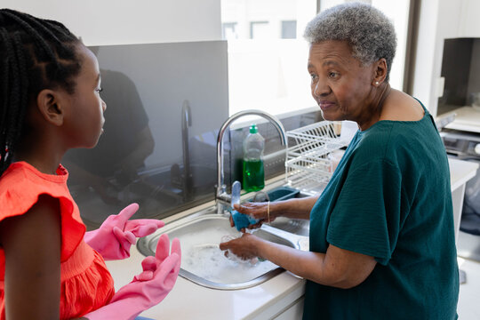 Senior female grandma and grandchild washing at kitchen sink, child in pink gloves with blue sponge