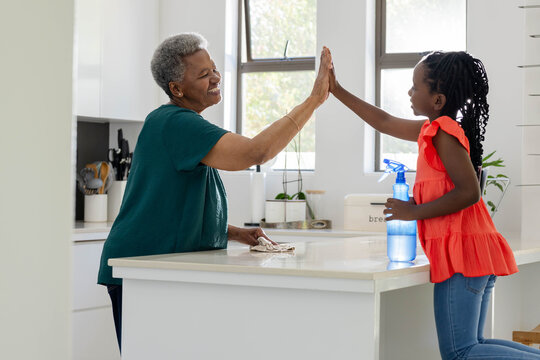 African American Senior woman, Youth girl high-fiving cleaning kitchen island, blue spray bottle