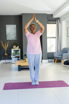 Senior African American woman doing standing yoga on purple mat at home wearing pink tee smartwatch
