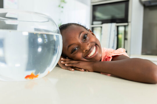 African American female child resting head on glossy countertop in kitchen watching goldfish bowl