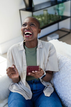 African woman laughing holding red smartphone sitting on sofa in bright living room with smartwatch