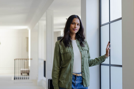 African American woman in 20s standing in hallway by window, hand on frame, wearing olive jacket