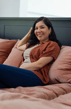 Female reclining on bed in bedroom wearing rust shirt and dark jeans with blush bedding