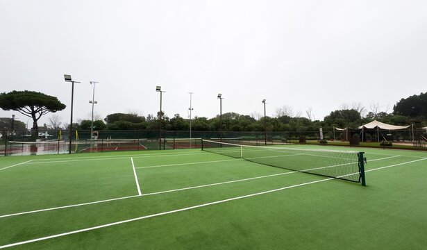Heavy rain pours down on empty tennis courts, soaking the surface and distorting the nets. The scene captures a sudden downpour disrupting play, with water splashing and reflecting the gloomy light.