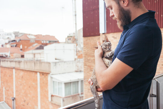 Young bearded man holding and cuddling his domestic cat on apartment balcony. Tender bonding moment between pet owner and feline in urban home setting. Concept of love for animals, companionship.