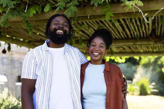 African American couple standing under wooden pergola with hanging vines, smiling at camera