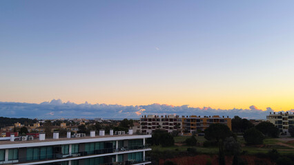 A softly blurred cityscape at sunset, with the sun partially obscured by buildings. Warm sunset colors dominate the sky, contrasting with the dark silhouett...