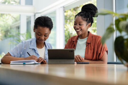 African adult and youth females sitting at wood table in study using tablet, notebook, pen, phone