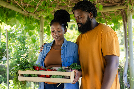 African couple holding wooden vegetable crate under pergola, woman in denim shirt, man in orange
